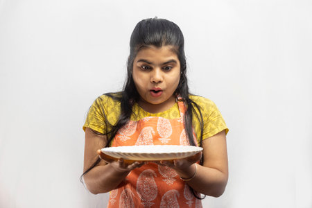 A pretty Indian housewife woman wearing cooking apron with a serving plate in hand looks down in surprise on white backgroundの写真素材