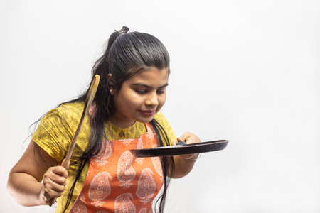 A pretty Indian housewife woman in cooking apron with wooden spatula and frying pan in hands on white backgroundの写真素材