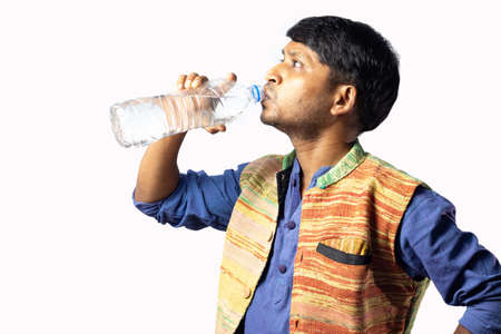 A young Indian male in ethnic dress drinks water from a disposable plastic bottle on white backgroundの写真素材
