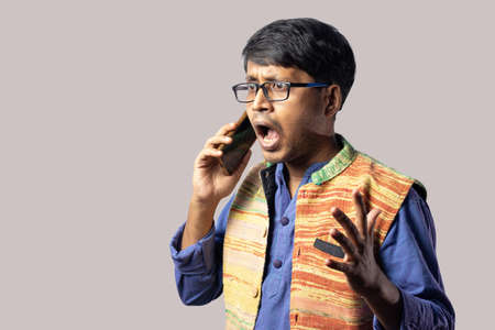 A young Indian male in blue dress talking over phone with agitated expression on gray background