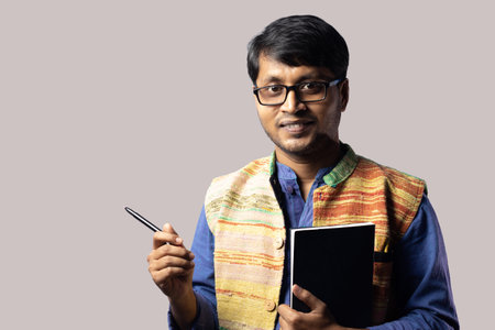 A young Indian male in ethnic dress standing with book and pointing with pen on gray backgroundの写真素材