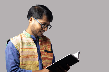 A young Indian male in ethnic dress reading book on gray background