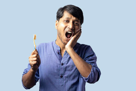 A young Indian male in blue dress facing teeth problem while brushing on blue backgroundの写真素材