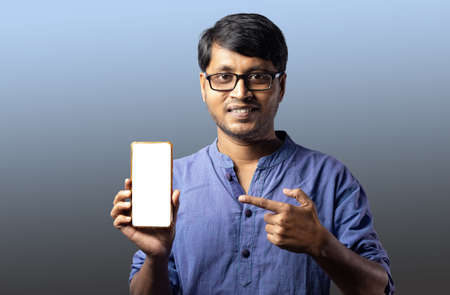 A young Indian male in blue dress pointing towards blank white screen of smart phone with smiling...