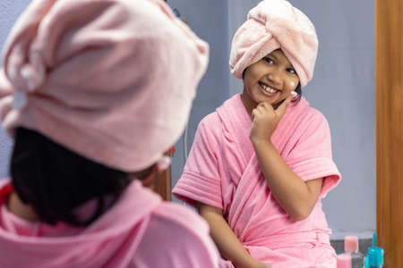A cute Indian girl child in pink bathrobe applying face cream in front of mirrorの写真素材