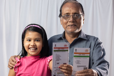 An Indian old man with his grand daughter smiling with Aadhaar card in hands on white background