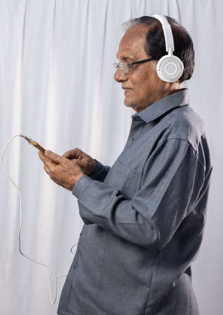 An Indian old person listening to music with smart phone and headphone on white backgroundの写真素材
