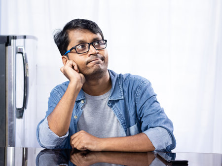 Portrait of a young middle aged Indian male wearing eyeglasses sitting at table and thinking upwards...