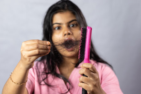 Selective focus on a pink hair brush full of fallen hair held in hand by a worried Indian woman on white backgroundの写真素材