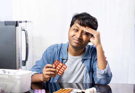 An Indian male with spectacles facing headache sitting beside a table with medicines on white backgroundの写真素材