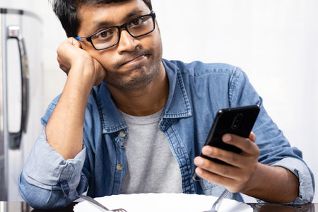 An Indian male waiting for food with smart phone sitting beside dining table on white background