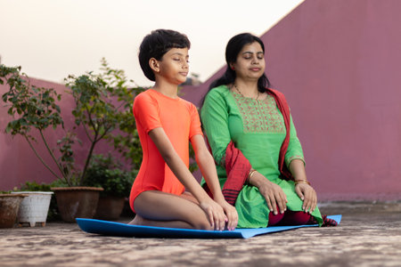 An Indian girl child with mother practicing Vajrasana yoga on yoga mat outdoorsの写真素材