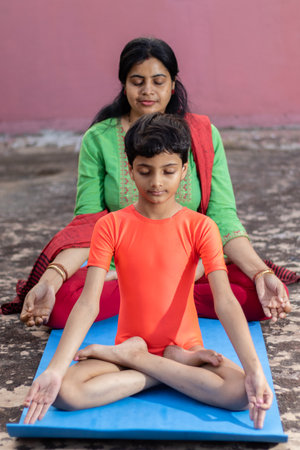 An Indian girl child with mother practicing padmasana yoga on yoga mat outdoorsの写真素材