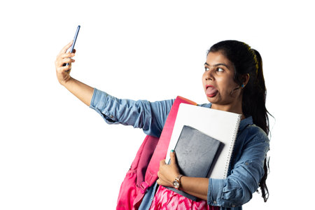 A pretty young Indian modern girl with books in hand taking selfie on white backgroundの写真素材