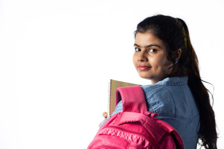 A pretty young Indian girl with books in hand and backpack looking back on white backgroundの写真素材