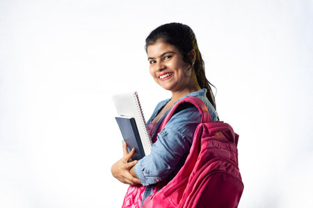 A pretty young Indian girl with books in hand and backpack standing with smiling face on white backgroundの写真素材