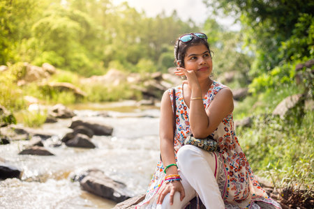 A pretty beautiful Indian woman sitting besides a rocky river in green backgroundの写真素材