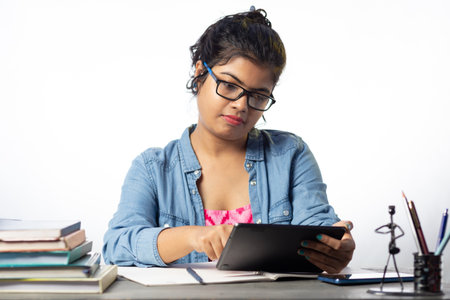 A pretty young Indian college student in eyeglasses studying with tablet on study table and white...