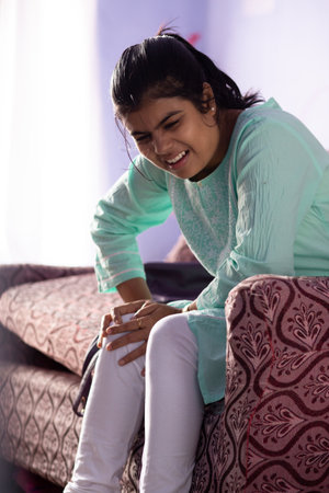 An Indian woman holding her knee in pain showing painful expression sitting on sofaの写真素材