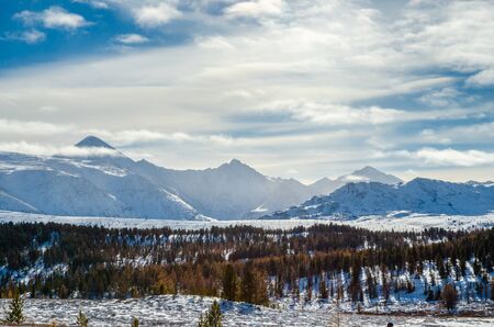 The autumn forest on the snow hills, far mountains and cloudsの写真素材