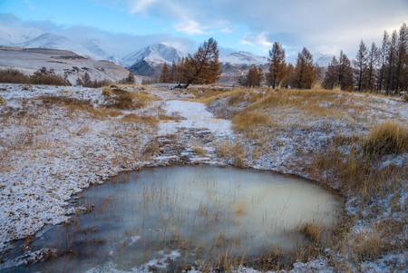 Ice on a pond and dry grass in a valley and a first snow in the mountainsの写真素材