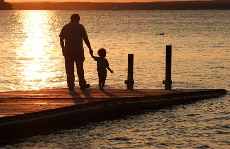 A father holds his son's hand as they walk out onto a dock at sunset.の写真素材