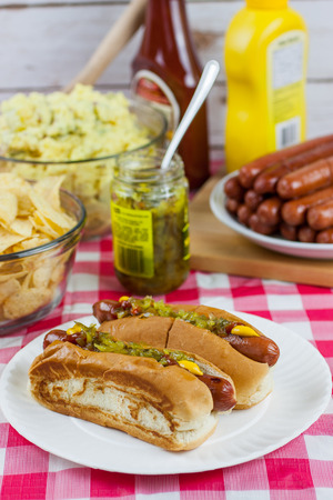 Grilled hot dogs on a paper plate sitting on a table with potato salad, chips, and condiments.の写真素材
