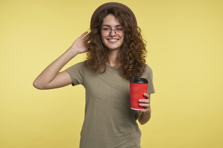 A studio shot of young student woman, she smiles and holding a coffee cup, wearing hat and eyeglasses, isolated over yellow backgroungの写真素材