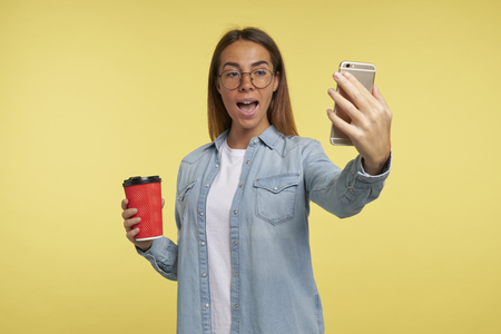 Pretty young woman makes a selfie on her smartphone, she holds a coffee cup, isolated over yellow backgroundの写真素材