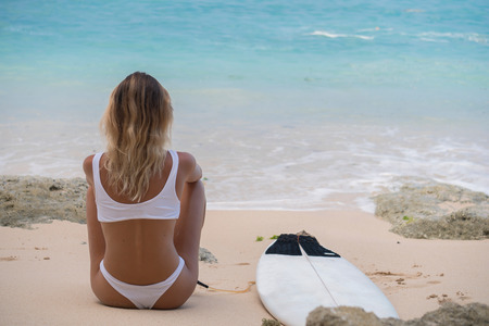 greatl back view of tanned bikini girl sitting on pink sand beach with surfboardの写真素材