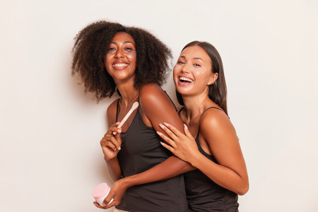 Two smiling young model girls in silk black tops standing body half-turned on a white colour...