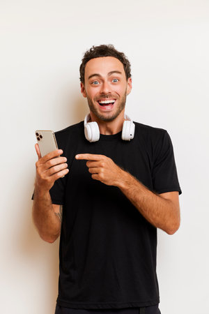 Young bearded guy smiles happily, pointing at his phone, dressed in black T-shirt, posing against a pale pink background, profit by the occasion concept, copy space, high quality photoの写真素材