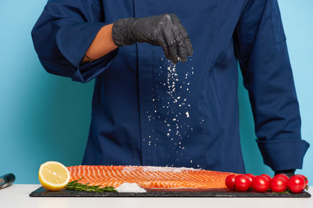 Photo of a man in blue chefs jacket and black gloves who salts fish fillet to make delicious meal, tasty food concept, copy space, high quality photoの写真素材