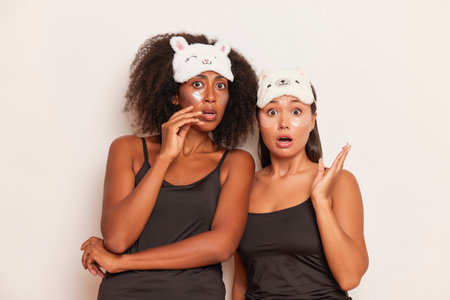 Two suprised young model girls dressed in silk black tops posing on a white colour background, they have speep masks on their heads, comfortable underclothing concept, copy space, high quality photoの写真素材