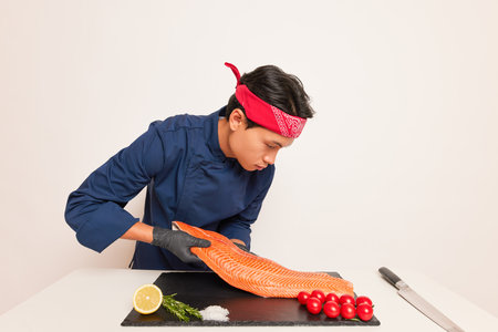 Young man in blue chefs clothes works with salmon fillet, asian chef is going to cook sushi and rolls, stands at the table with tomatoes and lemon, delicious food concept, copy space, high quality photoの写真素材