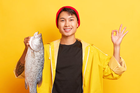 Happy guy in red cap and yellow jacket holding big fish in his hand, showing Ok sign with another...