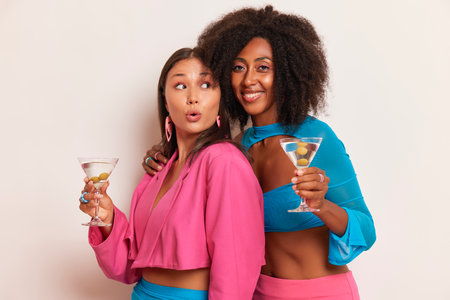 Two beautiful girl friends are in the studio, posing together with glass of martini, dressed in pink and blue clothes having fun, happy time concept, copy space, high quality photoの写真素材