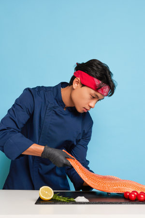 Young Asian cook posing on the blue studio background, wearing blue chefs jacket and gloves, examining the fish fillet for tasty sishi, natural food concept, copy space, high quality photoの写真素材