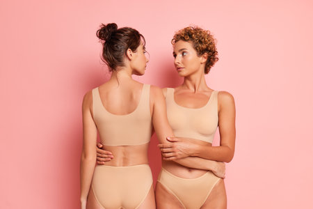Two half-naked girls are standing in the studio dressed only in panties and bras, looking at each other, one is facing the camera, other facing away, female friendship concept, copy space, high quality photoの写真素材