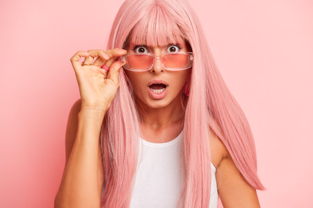 Surprised pretty girl in pink wig and stylish pink sunglasses poses against studio background, very surprised, lowering her glasses with one hand, interesting case concept, copy space, high quality photoの写真素材