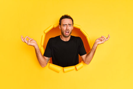 A young bearded guy stands in the middle of a hole in a yellow background, wears a black T-shirt, is surprised by something shrugs his shoulders and raises his arms to the sides, high quality photoの写真素材
