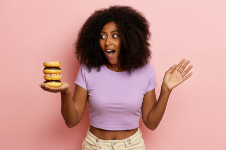 Portrait shot of surprised dark skinned young woman with overjoyed smile, keeps delicious doughnuts and looks excited, isolated over pink background. High quality photoの写真素材