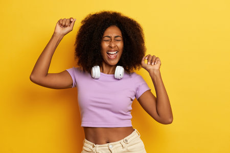 Horizontal shot of cheerful curly haired woman with hands raised up in the air, enjoys her dance in...