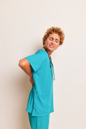 Health worker with curly hair in blue medical suit poses inside holds hands on her back, looks looks...