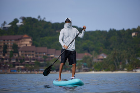 Guy in white hoodie and black shorts standing on board with paddle in hands, floating withf tropical coast on background, searching for wave, happy holiday concept, copy spaceの写真素材