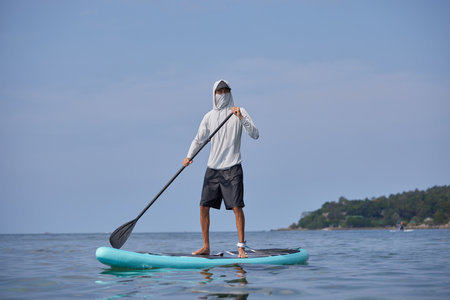 Surfer in white hoodie with mask on his face floating on surf board with oar in hands, standing on tropical coast background, sport life concept, copy spaceの写真素材