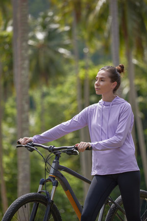 Young sporty woman in grey hoodie holds the steering wheel of his bicycle standing on palm trees on background, sport lifestyle concept, copy spaceの写真素材