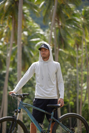 Young bearded man in white hoodie and black shorts stands next to his bicycle, holds the steering wheel and cycle seat. sport lifestyle concept, copy spaceの写真素材