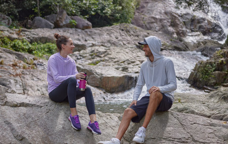 Couple of happy tourists having rest on the stone near mountain stream, girl holding bottle of water in her hands, guy looking at her and smiling, freedom lifestyle concept, copy spaceの写真素材