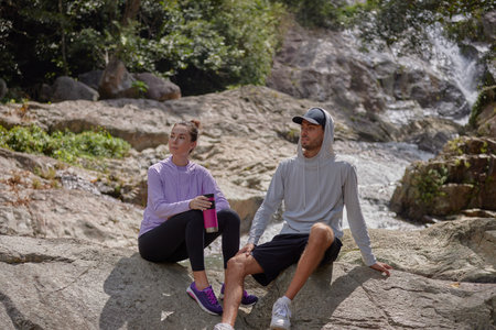 Couple of tourists resting near mountain stream, the girl holding bottle of water in her hands, the guy sitting leaning back on his hands, sport lifestyle concept, copy spaceの写真素材